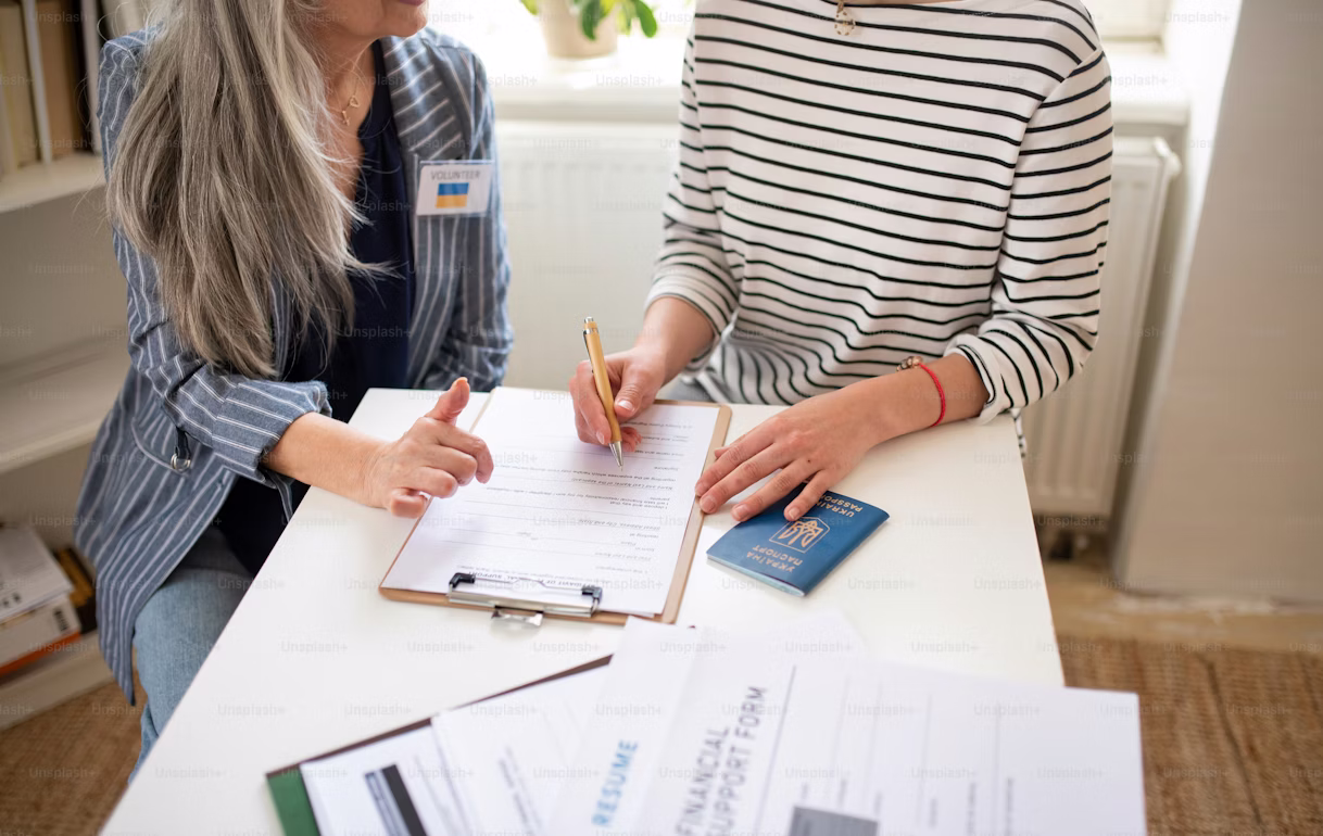 A volunteer assists a person in completing paperwork at a desk. A passport, a financial support form, and a resume are visible on the table.